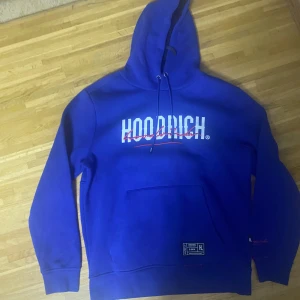 Blå hoodie från Hoodrich med tryck - Snygg blå hoodie från Hoodrich med stort vitt och rött tryck på bröstet och ryggen. Tröjan har huva med snörning, känguruficka och ribbade muddar. Perfekt för dig som gillar streetwear och vill sticka ut med en bold look.