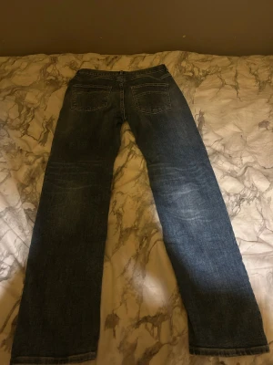 Blå raka jeans  - Säljer ett par klassiska blå jeans med rak passform och fyra fickor. Jeansen har en mörk tvätt med lätt slitning och är tillverkade i robust denim. Perfekt för dig som gillar en tidlös och enkel stil.