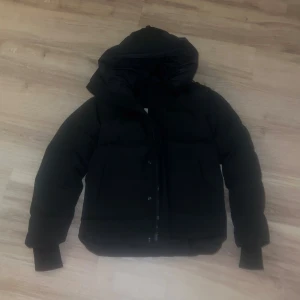 Svart pufferjacka från Canada Goose Maximillian  - Säljer en svart pufferjacka från Canada Goose med huva och ribbade muddar. Jackan har en snygg patch på ärmen och dragkedja framtill. Perfekt för dig som vill ha en varm och stilren jacka med cool streetkänsla.