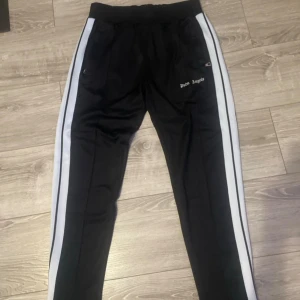 Palm Angels svarta mjukisbyxor med vita ränder - Svarta mjukisbyxor från Palm Angels med vita och svarta ränder längs sidorna. Byxorna har dragkedjor vid fickor och benslut, samt elastisk midja. Snyggt broderad Palm Angels-logga på framsidan. Perfekt för en streetwear-look.