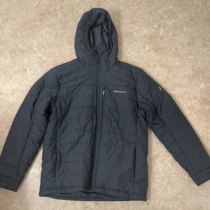 Svart dunjacka Peak Performance - Svart dunjacka från Peak Performance med huva och diskret logga på bröstet. Jackan har quiltad design, dragkedja framtill och en ficka med dragkedja. Perfekt för kalla dagar och har en clean, sportig look.