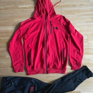 Röd vindjacka från The North Face - Snygg röd vindjacka från The North Face med huva och hel dragkedja framtill. Jackan har svarta detaljer, tre praktiska fickor med dragkedja och logga på bröstet. Perfekt för dig som gillar sportig stil och vill ha något som sticker ut.