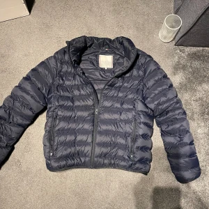 Mörkblå pufferjacka Tommy Hilfiger - Snygg mörkblå pufferjacka från Tommy Hilfiger med quiltad design och dragkedja framtill. Jackan har två sidofickor med dragkedja, hög krage med logotyp och diskret Hilfiger-flagga på bröstet nypris 2000 passar från 167 till 176