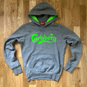 Grå Carlsberg hoodie med neontryck - Snygg grå hoodie från Carlsberg med stort neon-grönt logotryck på bröstet och matchande grön insida i huvan. Tröjan har klassisk känguruficka, ribbade muddar och mjukt bomullsmaterial. Står storlek M men passar mer som en S enligt mig.