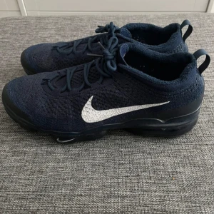 Nike Vapormax Flyknit marinblå sneakers - Säljer ett par Nike Vapormax Flyknit sneakers i marinblått med vit swoosh. Skorna har stickad ovandel i flyknit-material, snörning och en unik svart sula med synliga luftbubblor. Perfekta för dig som gillar sportig och modern stil.