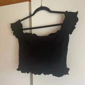 Svart smockad croppad topp - Säljer en svart smockad croppad topp med volangdetaljer vid ärmslut och nederkant. Toppen har en fyrkantig ringning och är ärmlös, perfekt för en trendig och avslappnad look. Storlek L men skulle säga mer S/M