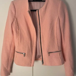 Rosa kavajjacka från Zara Woman - Snygg ljusrosa kavajjacka från Zara Woman med råa kanter och silverfärgade dragkedjor fram och vid fickorna. Jackan är tillverkad i ett strukturerat tyg och har lång ärm samt rund halsringning. Perfekt för dig som gillar stilrena detaljer.