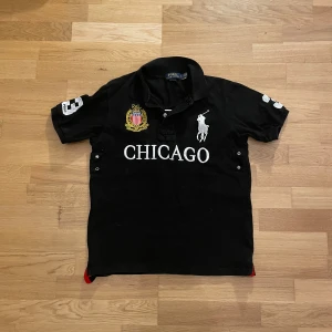Svart Polo Ralph Lauren polo Chicago - Svart t-shirt från Polo Ralph Lauren med broderad logga, stor CHICAGO-text och emblem framtill. Nummer 3 på ryggen och ärmen. Storlek L men passar bättre som storlek M