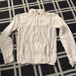 Vit vindjacka från A-Z - Säljer en stilren vit vindjacka från A-Z med hel dragkedja framtill och två fickor fram och en på ryggen. Jackan har lång ärm, hög krage, inbyggd luva vid kragen och svart detalj på ryggen. Perfekt för dig som gillar sportig och clean look.