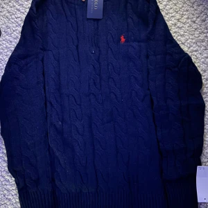 Marinblå kabelstickad kofta Polo Ralph Lauren - Marinblå kabelstickad kofta från Polo Ralph Lauren med röd broderad logga på bröstet. Tröjan har ribbade muddar, dragkedja framtill med läderdetalj och är långärmad. Perfekt för lager-på-lager och snygg till jeans.