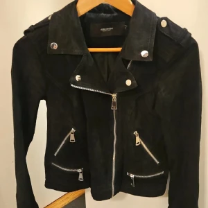 Svart moocajacka från Vero Moda - Snygg svart moocajacka från Vero Moda med flera silvriga dragkedjor och detaljer. Jackan har klassisk bikerstil med bred krage och långa ärmar. Perfekt för dig som vill ha en cool och tidlös look.