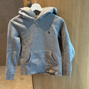 Grå hoodie från Polo Ralph Lauren - Snygg grå hoodie från Polo Ralph Lauren med klassisk huva och magficka. Tröjan har ett mörkblått broderat logomärke på bröstet och är tillverkad i mjuk bomull. 