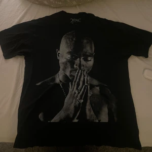 Svart Tupac t-shirt med tryck - Svart t-shirt från 2PAC med stort tryck av Tupac Shakur på framsidan och citat på baksidan. Klassisk rund hals och korta ärmar. Materialet känns som mjuk bomull, perfekt för dig som gillar streetwear och ikoniska prints.