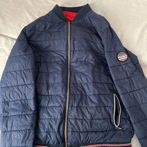 Mörkblå quiltad pufferjacka Jack & Jones - Snygg mörkblå pufferjacka från Jack & Jones med quiltad design och röd insida. Jackan har dragkedja framtill, ribbad krage och muddar samt en patch på ärmen. Perfekt för kyliga dagar och har en sportig vibe.