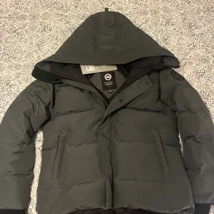 Mörkgrå dunjacka från Canada Goose black label  - Säljer en mörkgrå dunjacka från Canada Goose med huva och svarta detaljer. Jackan har en klassisk pufferstil, raka linjer och diskret logga på ärmen. Fodrad för extra värme och med dragkedja samt knappar framtill. Perfekt för kalla vinterdagar.