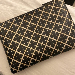 Svart och beige kuvertväska By Malene Birger - Kuvertväska från By Malene Birger med ikoniskt grafiskt mönster i svart och beige. Väskan har en rektangulär form, guldfärgad dragkedja och är tillverkad i skinn med textilfoder. Perfekt storlek för att bära det viktigaste och snygg att hålla i handen.