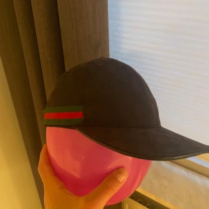 Svart Gucci keps  - Svart keps från Gucci med diskret GG-mönster över hela ytan och klassiska gröna och röda ränder på sidan. Tillverkad i Italien och gjord av bomull. Snygg och stilren design som ger en lyxig vibe till din outfit. Den är lite solbränd men man kan enkelt färglägga den.  