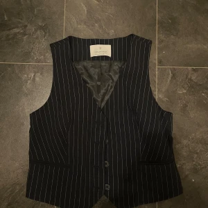 Randig svart väst från Stradivarius - Snygg svart väst från Stradivarius med tunna vita ränder. Klassisk passform med knappar framtill, två fickor och fodrad insida. Perfekt för att ge din outfit en stilren och trendig touch.