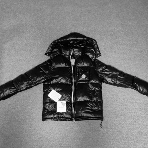 Svart Moncler pufferjacka med huva - Säljer en svart pufferjacka från Moncler med glansig finish och huva. Jackan har dragkedja, fickor med knappar och Moncler-logga på bröstet. Insidan har ett coolt serietryck. Perfekt för dig som vill ha en varm och snygg jacka med streetkänsla.