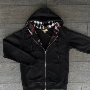 Svart Burberry hoodie med dragkedja - Svart hoodie från Burberry med klassisk rutmönstrad insida i huvan med vita, grå och röda detaljer. Tröjan har huva med snörning, två fickor fram och hel dragkedja. Perfekt för dig som gillar stilrena streetwear-plagg med lyxig touch.