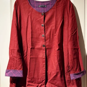 Rött set från Gudrun Sjödén - Säljer ett vävt rött set från Gudrun Sjödén med kavaj och byxa. Kavajen har knappar och lila-rutigt foder som syns vid ärmslut och krage. Byxorna är vida.. kläderna är i fint skick!