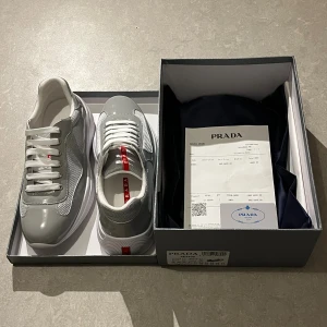 Prada America’s Cup Original Sneakers - Snygga gråa sneakers från Prada med vita detaljer och röda loggor på plösen och sulan. Skorna har en mix av glansigt skinn och mesh, vita snören och en chunky vit sula. Perfekta för dig som vill ha en stilren och exklusiv look. Pris kan diskuteras!