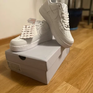 Nike Air Force 1 '07 helvita sneakers - Nike Air Force 1 '07 i helvitt skinn med klassisk siluett, perforerad tå och vit sula. Snygga detaljer som AF1-tag på snörningen och ikonisk Swoosh på sidorna. Perfekta för dig som vill ha en clean och tidlös sneaker-look.