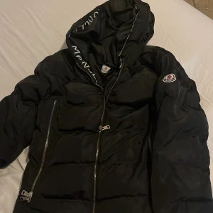 Svart pufferjacka från Moncler - Säljer en svart pufferjacka från Moncler med huva och logotyp på ärmen. Jackan har två sidofickor med dragkedja och en snygg Moncler-text på huvan. Perfekt för kalla dagar och riktigt stilren look.