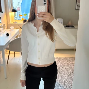 Vit kofta med knappar - Säljer en vit kofta med långa ärmar och knappar framtill. Tröjan har en mjuk ribbad kant vid midjan och är tillverkad i ett tunt, bekvämt material. 