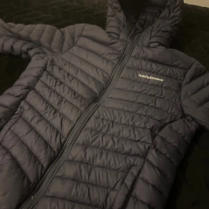 Mörkblå Peak Performance dunjacka - Snygg mörkblå dunjacka från Peak Performance med huva och hel dragkedja. Jackan har ett quiltat mönster och logga på bröstet. Perfekt för kyliga dagar och har en modern, normal passform. Materialet är lätt och syntetiskt, vilket gör den smidig att bära.