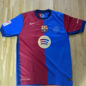 FC Barcelona Lamine Yamal matchtröja - Säljer en officiell FC Barcelona fotbollströja från Nike med Lamine Yamal och nummer 19 på ryggen. Tröjan har korta ärmar, är delad i röd och blå med klubbmärke, Spotify-logga och UNHCR-tryck. Materialet är lätt polyester, perfekt för match eller träning.