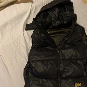 Svart dunväst från Black Good - Svart dunväst från Black Good med avtagbar huva och dragkedja framtill. Västen har en quiltad design och är tillverkad i glansig polyester. Perfekt för att hålla värmen under kyliga dagar och har en modern, sportig look.