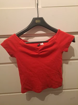 Röd offshoulder topp från H&M Divided - Säljer en röd offshoulder topp från H&M Divided i storlek S. Toppen är ribbad, har korta ärmar och en snygg rynkdetalj vid bysten. Perfekt för dig som gillar en croppad och trendig stil.