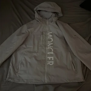 Vvindjacka från Moncler med huva - Snygg grå vindjacka från Moncler med stor logga framtill, huva och dragkedjor på fickorna. Jackan har en clean look med diskret Moncler-märke på ärmen och en liten detalj i rött, vitt och blått på ryggen. Perfekt för dig som gillar stilrena och exklusiva plagg. Passar L också 