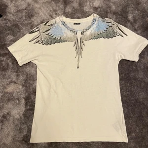 Marcelo Burlon vit t-shirt med vingar - Säljer en vit t-shirt från Marcelo Burlon med ikoniskt vingtryck i svart och blått runt halsen. T-shirten är i 100% bomull och har en normal passform med korta ärmar. Perfekt statement-plagg för dig som gillar unik design.