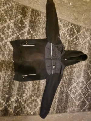 Svart fleecejacka med huva från Lacoste - Svart fleecejacka från Lacoste i storlek S. Jackan har huva, hel dragkedja och två fickor med dragkedja. Paneler i syntetmaterial på bröstet och detaljer med Lacoste-logga. Perfekt för dig som gillar sportig och clean stil.