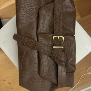 Brun kuvertväska i krokopräglat skinn - Snygg brun kuvertväska i krokopräglat skinnimitation med guldfärgat spänne. Väskan har en stilren design och ett bälte som detalj framtill. Perfekt accessoar för att lyfta din outfit.