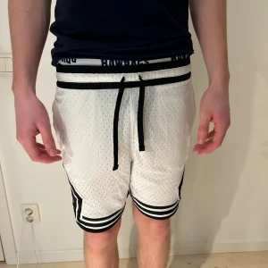 Jordan vita basketshorts med snörning - Snygga vita Jordan basketshorts med svarta detaljer och snörning i midjan. Shortsen har perforerat meshmaterial för extra ventilation och breda svarta ränder längst ner. Ikonisk Jumpman-logga på sidan och Dri-Fit-tryck vid midjan. Perfekta för sport och chill.