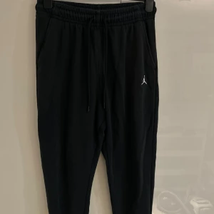Svarta Jordan mjukisbyxor med snörning - Svarta mjukisbyxor från Jordan med broderad Jumpman-logga i vitt på benet. Byxorna har elastisk midja med snörning, ribbade muddar vid benslut och mjukt fleecefoder på insidan. Perfekta för chill eller träning.
