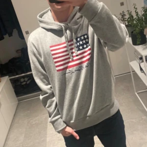 Grå hoodie Polo Ralph Lauren M - Snygg grå hoodie från Polo Ralph Lauren i storlek M. Framsidan har ett stort tryck med amerikanska flaggan i rött, vitt och blått samt märkets signatur under. Hoodien har snörning vid huvan och är tillverkad i mjuk bomullsmix. Perfekt för chill dagar.