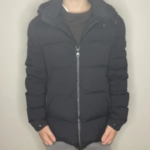 Svart dunjacka från J.Lindeberg - Svart dunjacka från J.Lindeberg med huva och snyggt emblem på ärmen. Jackan har en rak passform, dragkedja framtill och två sidofickor med zip. Modellen är 176 och väger ca 68kg. Finns ett litet defekt i en av fickorna men syns inte vid användning!