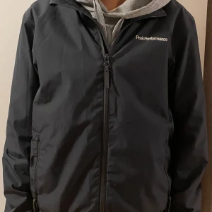 Peak Performance Jacka - Svart vindjacka från Peak Performance med stilren design och diskret logga på bröstet. Jackan har dragkedja framtill, två sidofickor med dragkedja och långärmade ärmar. Materialet är syntet och jackan har en normal passform.