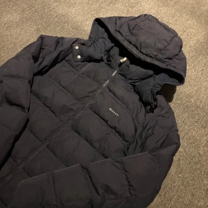 Mörkblå pufferjacka från GANT - Snygg mörkblå pufferjacka från GANT med huva och dragkedja framtill. Jackan har quiltad design och logga på bröstet. Perfekt för kalla dagar och har en stilren look som funkar till det mesta. Huva med tryckknappar och långa ärmar. Storlek: 176-XS