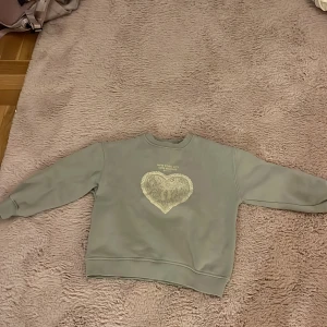 Grå/blå sweatshirt med hjärtmotiv - Grå sweatshirt från Zara med tryck framtill som visar ett stort hjärta med spetsdetaljer och rosett. Texten 'New York City Cute Friends Est. 1987' ovanför motivet. Tröjan har rund hals och långa ärmar med ribbade muddar. Perfekt för chill dagar.