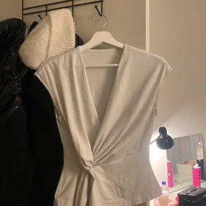 Vit omlottopp med knyt i midjan - Säljer en vit omlottopp med djup v-ringning och knyt i midjan. Toppen är ärmlös och har en snygg, figurnära passform som framhäver midjan. Perfekt att styla med jeans eller kjol för en clean look.