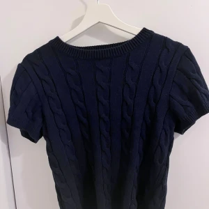 Mörkblå stickad tröja Gina Tricot - Snygg mörkblå stickad tröja från Gina Tricot med korta ärmar och grovt kabelstickat mönster. Rund halsringning med ribbad kant och mjukt material som känns skönt mot huden. Perfekt för dig som gillar klassisk stil med en modern twist. Aldrig använd, endast testad.