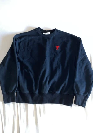 Svart sweatshirt från Ami med rött hjärta - Svart sweatshirt från Ami Alexandre Mattiussi med ett broderat rött hjärta och A på bröstet. Tröjan har rund hals, ribbade muddar och är långärmad. Tillverkad i mjuk bomull, perfekt för en chill och stilren look.