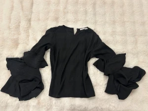 Svart blus med volangärmar från NA-KD - Supersnygg svart blus från NA-KD med dramatiska volangärmar och lätt v-ringning. Toppens material har en lätt glans och känns flowy. Perfekt för dig som vill sticka ut med coola detaljer och en trendig siluett.