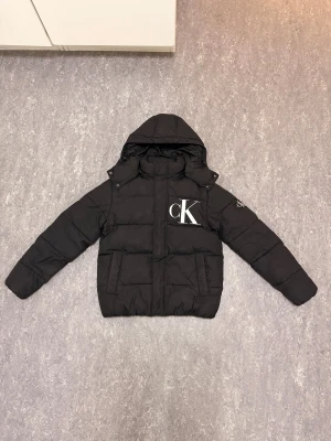 Svart pufferjacka från Calvin Klein - Svart pufferjacka från Calvin Klein Jeans med stor CK-logga på bröstet och huva. Jackan har långa ärmar, två fickor fram och quiltad design. Perfekt för kalla dagar och har en clean, modern look som passar streetstyle.