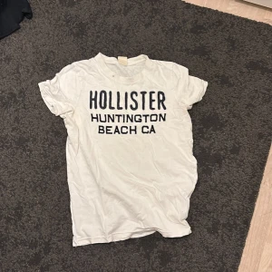 Vit Hollister t-shirt med tryck - Vit t-shirt från Hollister med svart text 'HOLLISTER HUNTINGTON BEACH CA' på bröstet. Klassisk rund halsringning och korta ärmar. Tillverkad i mjuk bomull, perfekt för en avslappnad och cool stil.